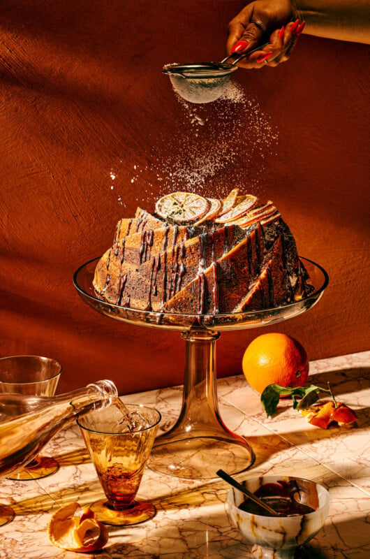 A marble cake garnished with lime and apple slices sits on a glass cake stand. A hand dusts it with sugar. A bottle pours liquid into a nearby glass. An orange and strawberries are on the marble surface, creating a vibrant still life scene.