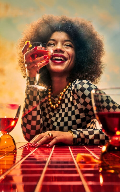 A woman with curly hair, wearing a checkered outfit and gold jewelry, smiles while holding a cocktail glass. She sits at a red tiled table with other glasses in front of her against a colorful background.