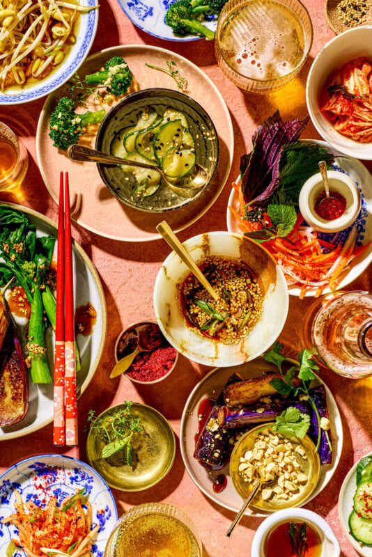 A vibrant display of various Asian dishes on a pink table. Plates are filled with pickles, kimchi, vegetables, spices, and garnishes. Red chopsticks and glasses add color and texture to the rich culinary spread.