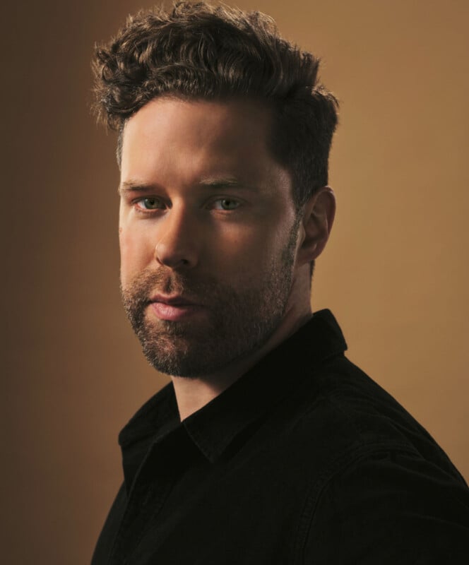 A man with short, curly hair and a beard is wearing a black shirt. He looks directly at the camera against a plain, warm-toned background.