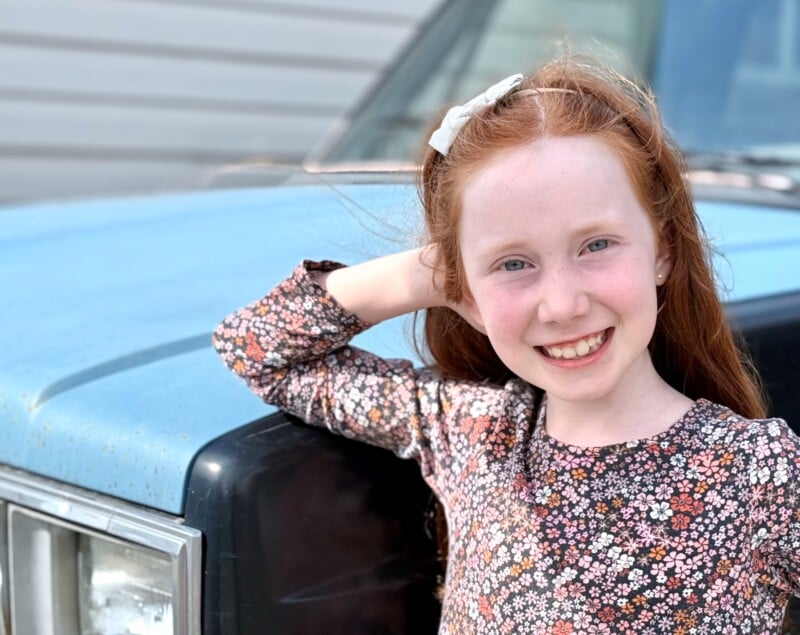 A young girl with long red hair, wearing a floral dress, smiles at the camera. She is standing next to an old blue truck with one hand resting on its hood. A building with horizontal siding is visible in the background.