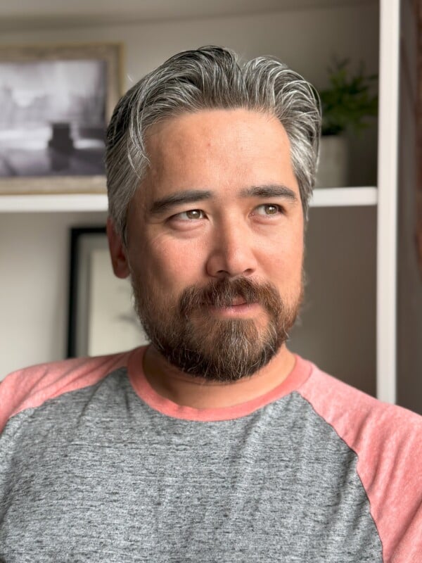 A person with salt and pepper hair and a beard gazes to the right in a thoughtful manner. They are wearing a gray and pink raglan t-shirt. The background includes shelves with a framed picture and a plant.