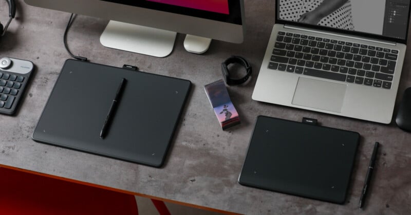 A workspace with a laptop, a desktop monitor, two drawing tablets with pens, a smartphone, a smartwatch, and a keyboard. The surface is a textured gray, and a red chair is partially visible.