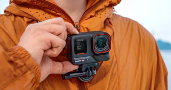 A person in a brown jacket holds an action camera close to their chest. The camera screen displays recording information. The background is blurred, focusing on the camera and the hand.