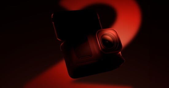 A compact camera with a rugged design is shown against a dark background with dramatic red lighting, featuring prominent shadows and highlights.