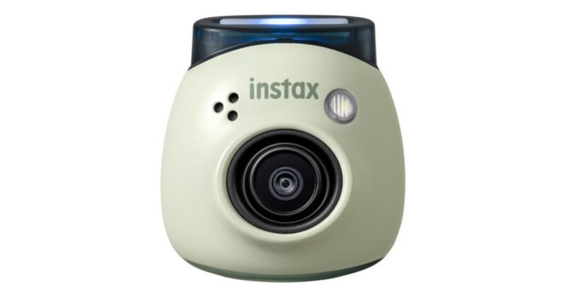 A compact, light-colored instant camera with a dark lens and "instax" branding on the front. The camera has a curved, cylindrical design and a thin, dark section at the top, possibly for a viewfinder or flash.