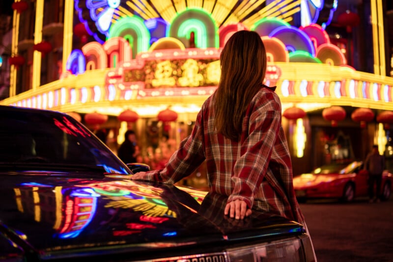 A person in a plaid shirt leans against a car, facing a vibrant neon-lit street with colorful signs and decorations. The scene is aglow with red, yellow, and green lights, creating a lively and bustling atmosphere.