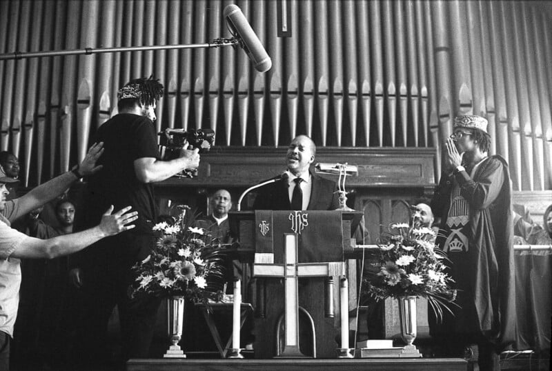 A black and white photo of a man speaking at a church pulpit, surrounded by a film crew and people in ceremonial attire. The background features large organ pipes and floral arrangements near the pulpit.