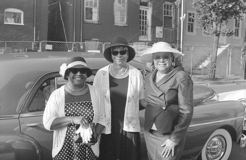 Three women in vintage attire and hats stand smiling in front of a classic car on a street with brick buildings. The black-and-white photo exudes a retro feel.