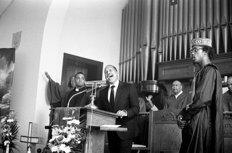 A man speaks passionately at a podium in a church, flanked by clergy and choir members. One wears a robe and another, a ceremonial hat. Flowers adorn the podium, and an organ is visible in the background.