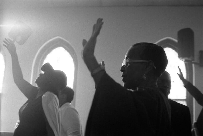 A monochrome image of people inside a church with hands raised, possibly during a worship service. Light filters through two stained glass windows, adding a serene atmosphere.