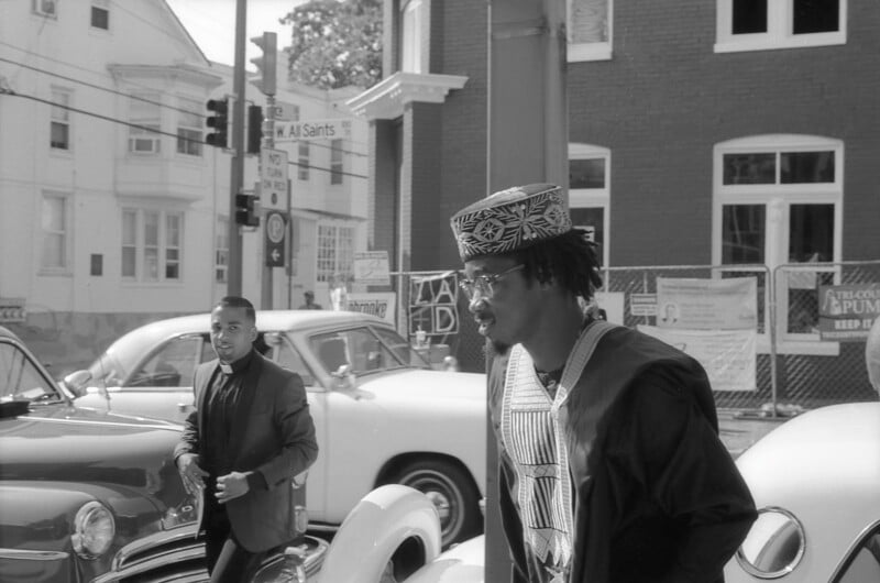 Two people are walking in a street with classic cars parked nearby. One of them is wearing a patterned hat and traditional garment, while the other is in a suit. A brick building and signs are visible in the background. The image is in black and white.