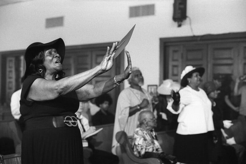 A group of people in a church setting, with a woman in the foreground passionately singing or speaking, wearing a hat and holding a paper. Several others in the background are also engaged in the service. The scene is in black and white.