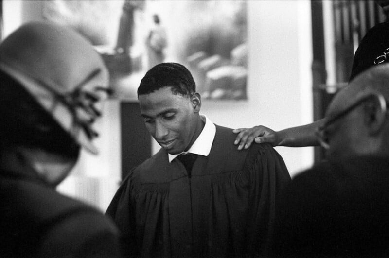 A black and white photo of a man in a robe standing with his head bowed. A hand rests on his shoulder, suggesting comfort or support. Other people are blurred in the foreground, creating a solemn and reflective atmosphere.