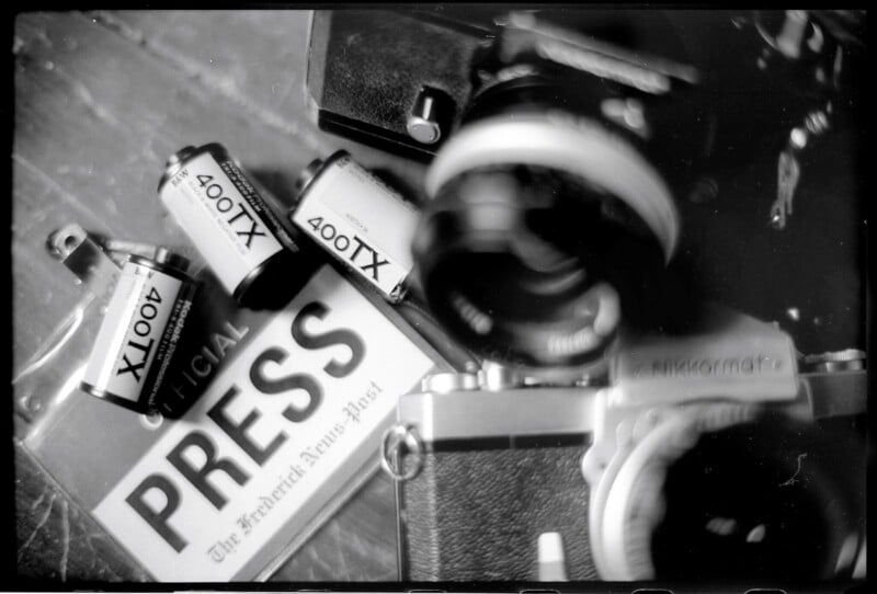 Black and white image of a vintage camera with a blurred lens in the foreground. Surrounding it are film rolls labeled "400TX" and a press badge from "The Frederick News-Post" lying on a wooden surface.