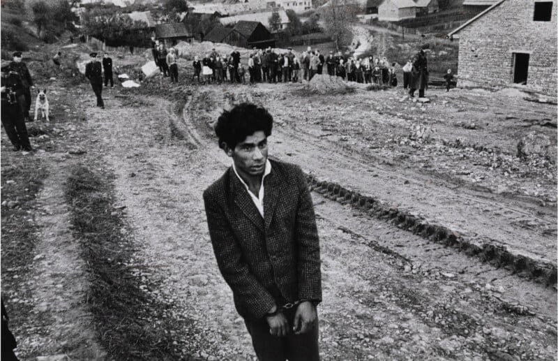 A young man stands on a dirt road, looking pensive. In the background, a group of people gather near some wooden houses. The scene conveys a rural, historic atmosphere.