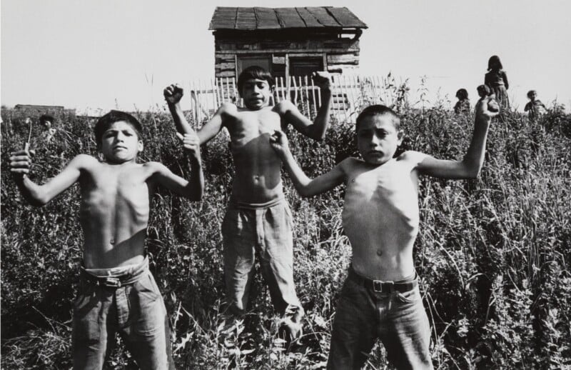 Three shirtless boys flex their muscles in a grassy field, with a small wooden shack in the background. A person stands near the shack. The scene is monochromatic.