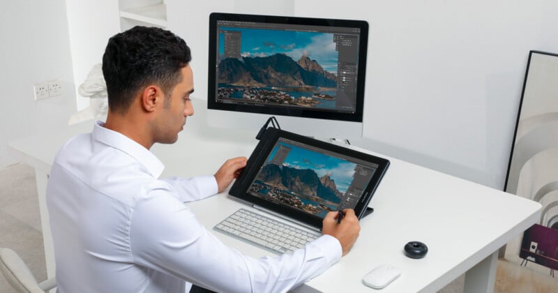 A man in a white shirt uses a stylus to edit a landscape photo on a graphics tablet, with a computer monitor displaying the same photo. He sits at a modern white desk with a keyboard, mouse, and some gadgets. The room is bright and minimalistic.