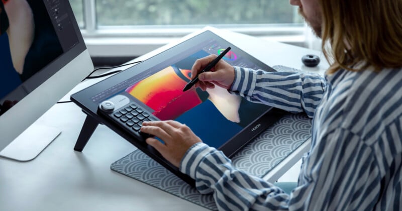 A person using a digital pen to draw on a graphic tablet, with vibrant colors on the screen. The setup includes a large monitor and a patterned desk mat. The person wears a striped shirt, and there's a window in the background.