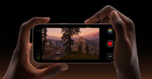 A person holds a smartphone horizontally, displaying a scenic view of a mountainous landscape with a road and a car at sunset. The phone screen shows camera controls.