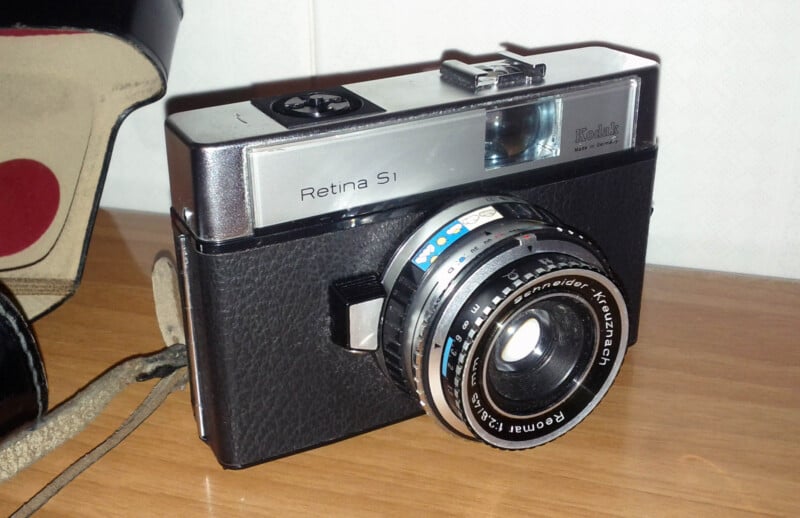 A vintage Kodak Retina S1 camera with a black textured body and silver accents, featuring a prominent lens. It is placed on a wooden surface next to an open leather case with a red interior lining.