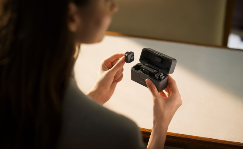 A person holding a black wireless earbud and its charging case. The case is open, showing one earbud inside. The background is a blurred table surface.