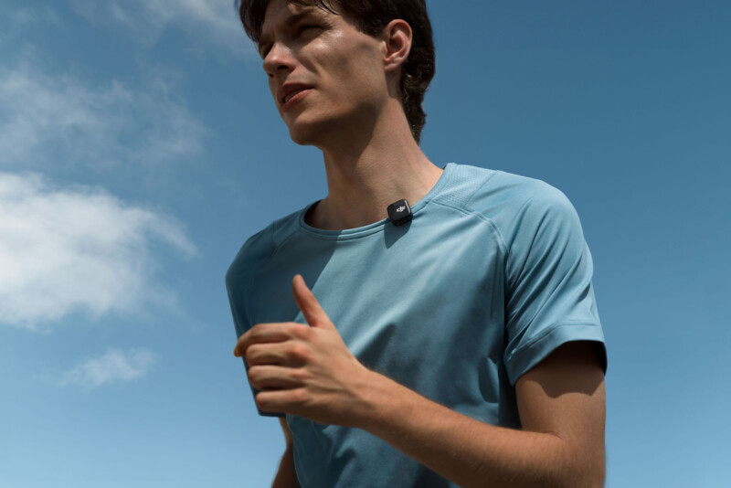 A person in a light blue shirt is jogging outdoors under a clear blue sky. They are wearing a small electronic device clipped to their shirt.