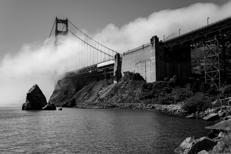 Black and white photo of the Golden Gate Bridge partially obscured by fog. The bridge extends over the water, connecting rugged terrain on either side. Vegetation and rocks line the shore in the foreground, with a large rock formation emerging from the water.