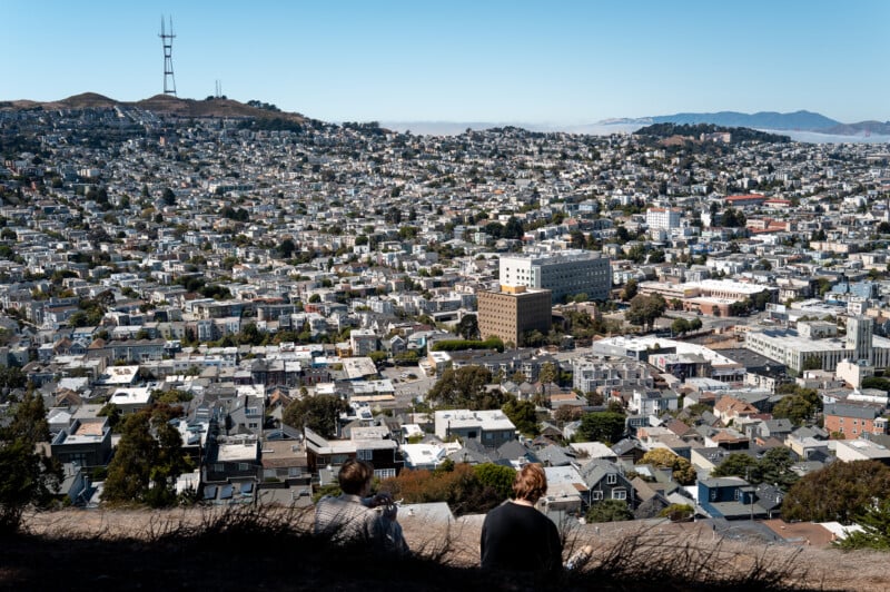 Two people sit on a hill overlooking a sprawling urban landscape on a clear day. The city is densely packed with buildings and houses. In the background, there are mountains and a tall radio tower atop a hill. The sky is blue and cloudless.