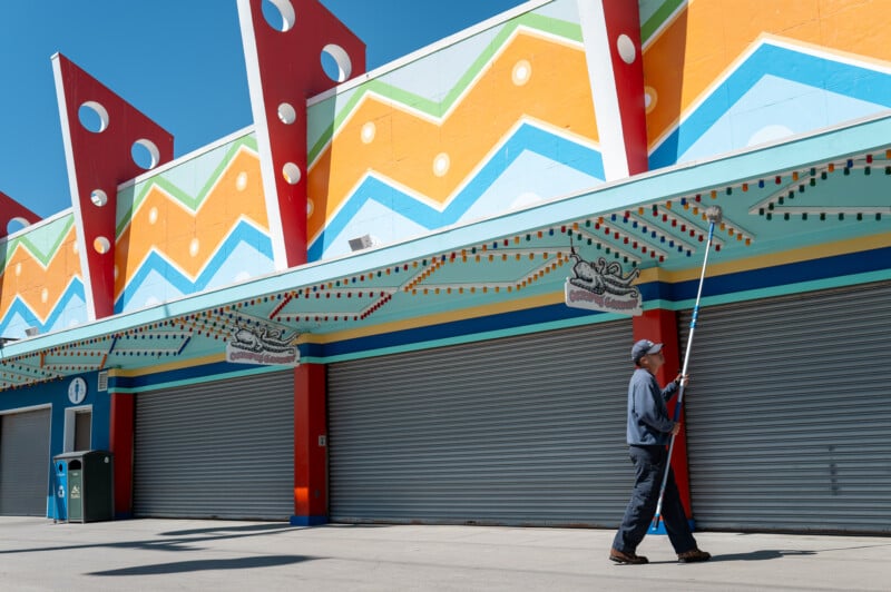 A person in casual attire uses a long pole to paint colorful patterns on the facade of a closed amusement park booth. The vibrant mural features zigzag shapes and geometric designs in blue, orange, yellow, and red. The day is sunny with a clear blue sky.