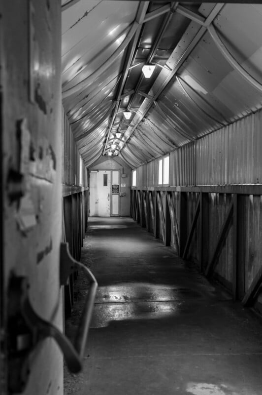 A black and white photo of a long, narrow, and dimly lit corridor with a corrugated metal roof and walls. The floor is wet, and a door is partially open on the left side. The end of the corridor features a closed door with a small window above it.