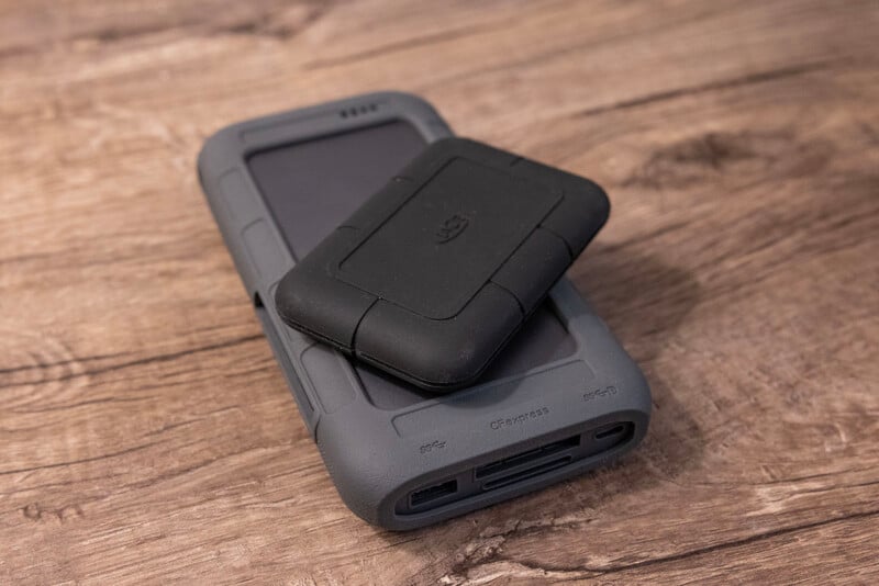 A rugged, black portable hard drive sits atop a larger, gray external battery pack on a wooden surface. The battery pack has USB and other ports visible at one end. Both devices appear durable with protective casings.