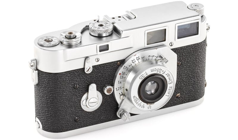 A vintage silver and black camera with textured grip and various dials on top. The lens has engraved markings and the body features a viewfinder and additional control knobs.