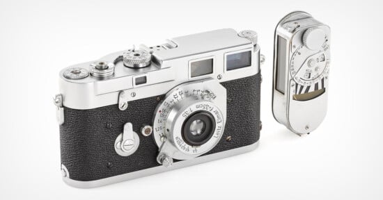 A vintage film camera with a black textured body and silver lens, accompanied by a compact, silver light meter. Both items are shown on a white surface against a neutral background.