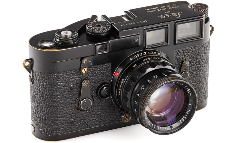 A vintage black Leica camera with textured grip and a large lens. The top displays various dials and markings, including "M3" and "919119." The lens has engraved details, and the body has a classic, elegant design.