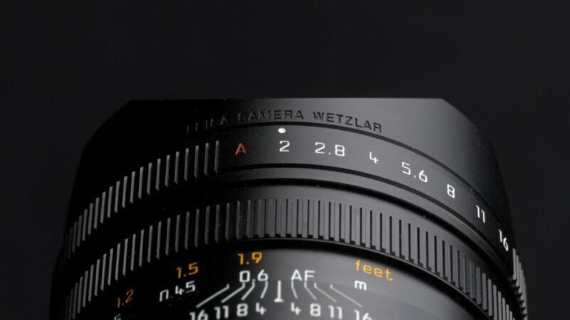 Close-up of a black camera lens focusing on the aperture settings, displaying f-stops from 2 to 16. The words "LEICA CAMERA WETZLAR" are inscribed at the top. The lens rings feature detailed distance markings in meters and feet.