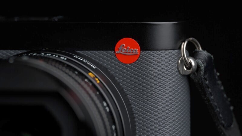 Close-up of a black Leica camera showing the textured surface, a portion of the lens, and the distinctive red Leica logo. The metallic strap eyelet is also visible on the right. The background is dark, emphasizing the camera's sleek design.