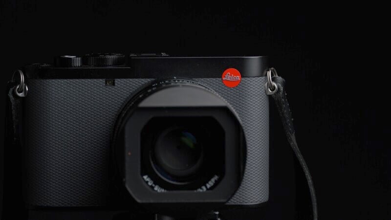 A professional black Leica camera with a prominent red logo is displayed against a dark background. The camera features a textured grip and is mounted on a tripod.