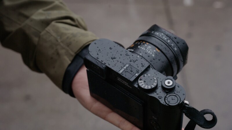 A person holding a black camera in their hand, covered with water droplets, indicating rainy weather. The person is wearing a green jacket, partially visible in the frame. The background is blurred, focusing attention on the camera.