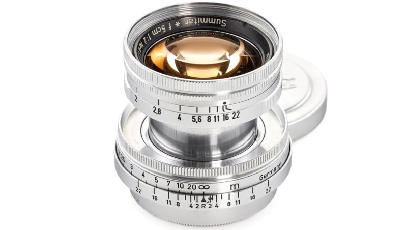 A shiny, silver camera lens with engraved markings and dials, showing a maximum aperture of f/2.8 and focus distances in meters. The lens cap is visible in the background.