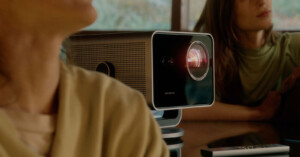 A close-up of a modern projector on a table, projecting light. Two individuals sit nearby, partially visible. A remote control rests on the table beside the projector. Blurred background with natural light coming through a window.