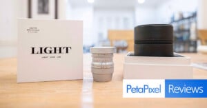 A photography lens on a table, accompanied by its packaging box and a storage case. The box features "LIGHT" branding and a small text, "Petapixel Reviews," is overlaid on the bottom right. The background is an out-of-focus room.