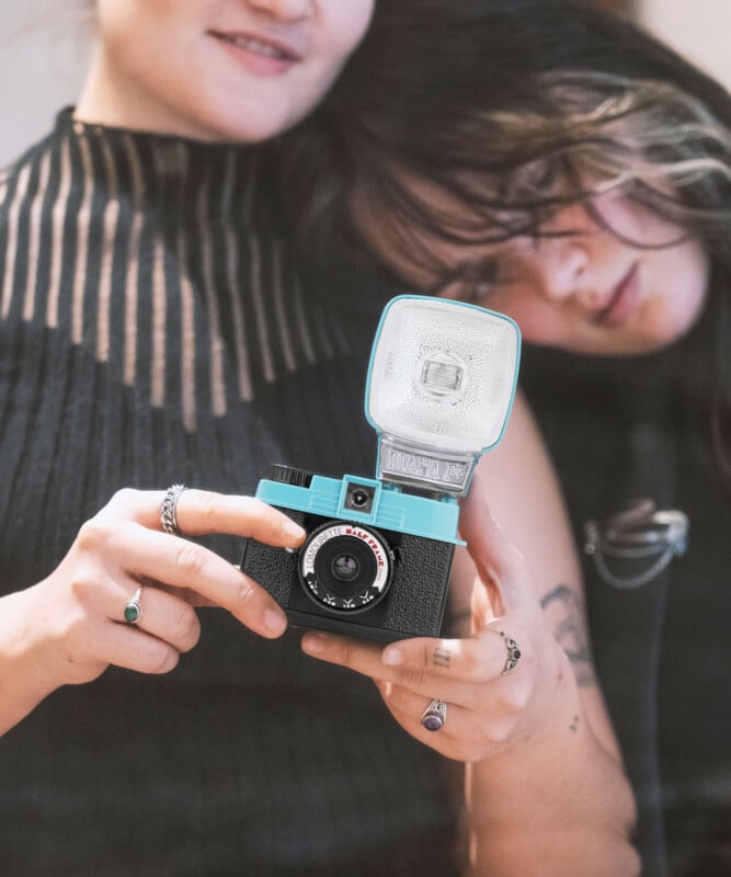 Two people are closely examining a vintage camera with a bright turquoise exterior and flash attachment. They both have long hair, and visible tattoos and rings adorn their hands.