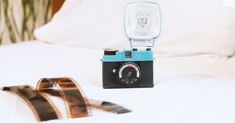 A vintage-style camera with a flash is placed on a white surface, next to two rolls of exposed film. The camera has a black body and a blue flash unit, adding a retro aesthetic to the scene.