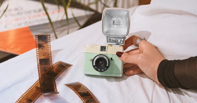 A person holds a vintage green camera with a silver flash attachment. Nearby, several strips of film negatives are spread out on a white cloth. In the background, partially visible plant leaves and blurred text can be seen.