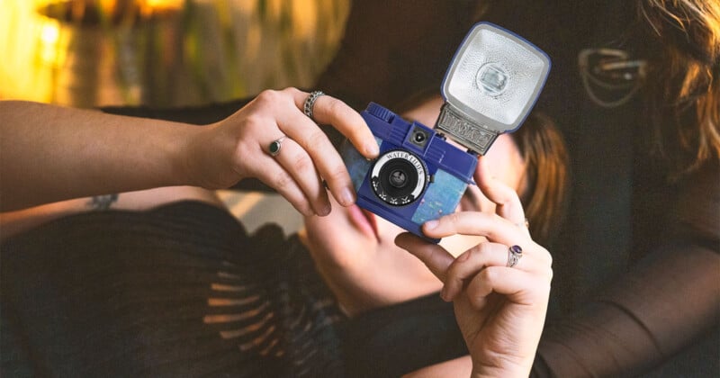 A person holding a vintage camera with a flash, wearing a black top and rings on their fingers. The background is softly lit, giving a warm ambiance.