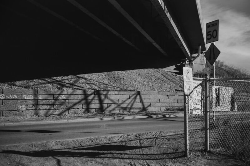 Black and white photo of a road under a bridge. A shadow of a structure is cast on a graffiti-covered retaining wall. A chain-link fence with signs and a speed limit sign of 50 km/h are visible. Sparse foliage is seen in the background.