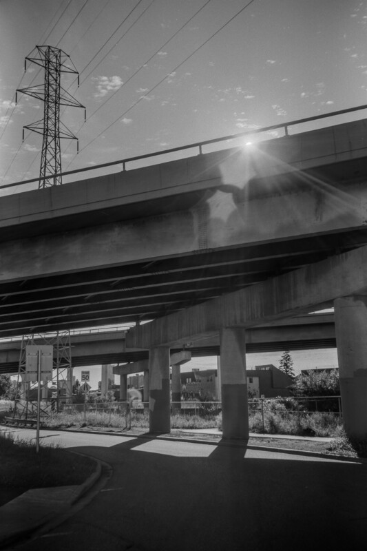 A black and white image of an overpass with sunlight peeking over the edge, casting shadows on the road below. A tall electrical tower stands nearby. The scene is urban with a partially cloudy sky.