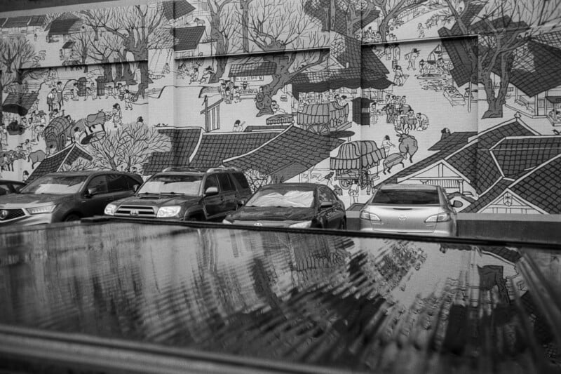 Black and white photo of several parked cars in front of a large mural depicting an intricate scene with people and traditional buildings. The mural and cars are reflected on a wet surface in the foreground.