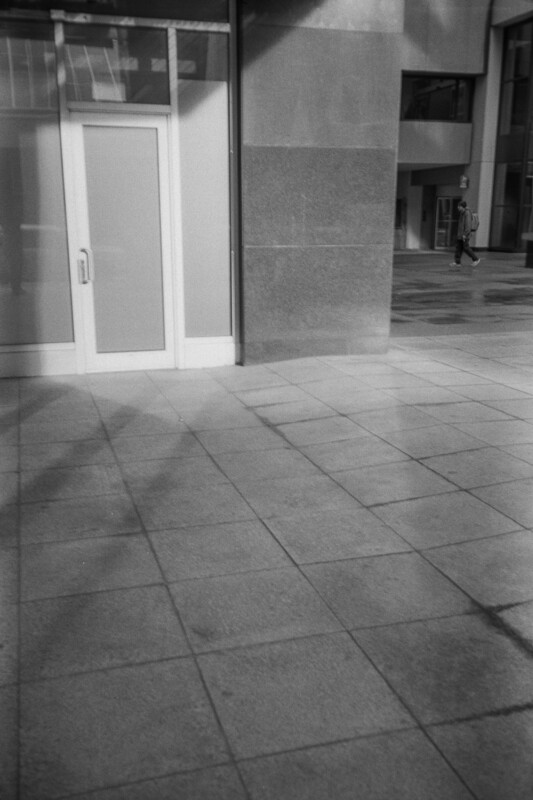 A black and white photo of a modern building entrance with a glass door. The sun casts shadows on the tiled pavement. In the background, a person walks across a courtyard near other buildings.